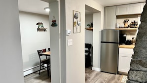 Una cocina compacta con refrigerador de acero inoxidable, una mesa de comedor de madera y sillas. Se observan interruptores de luz en la pared y un pequeño estante con utensilios de cocina.