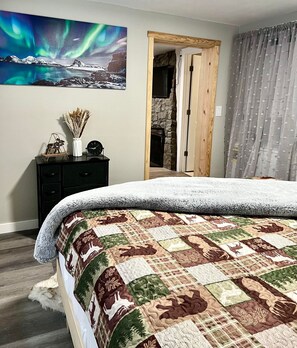 A bedroom with a large bed, a painting of the Northern Lights, a dresser, and a stone fireplace. - Log cabin in heart of Anchorage (Anchorage)