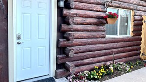Una cabaña de troncos con una puerta blanca, una ventana con contraventanas y un macizo de flores variado.