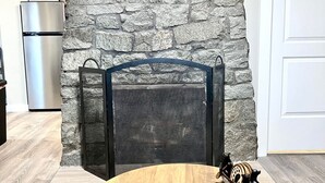Una sala de estar con pared de piedra, televisor montado, chimenea con puerta de cristal, mesa de madera redonda y dos controles remotos sobre la mesa.