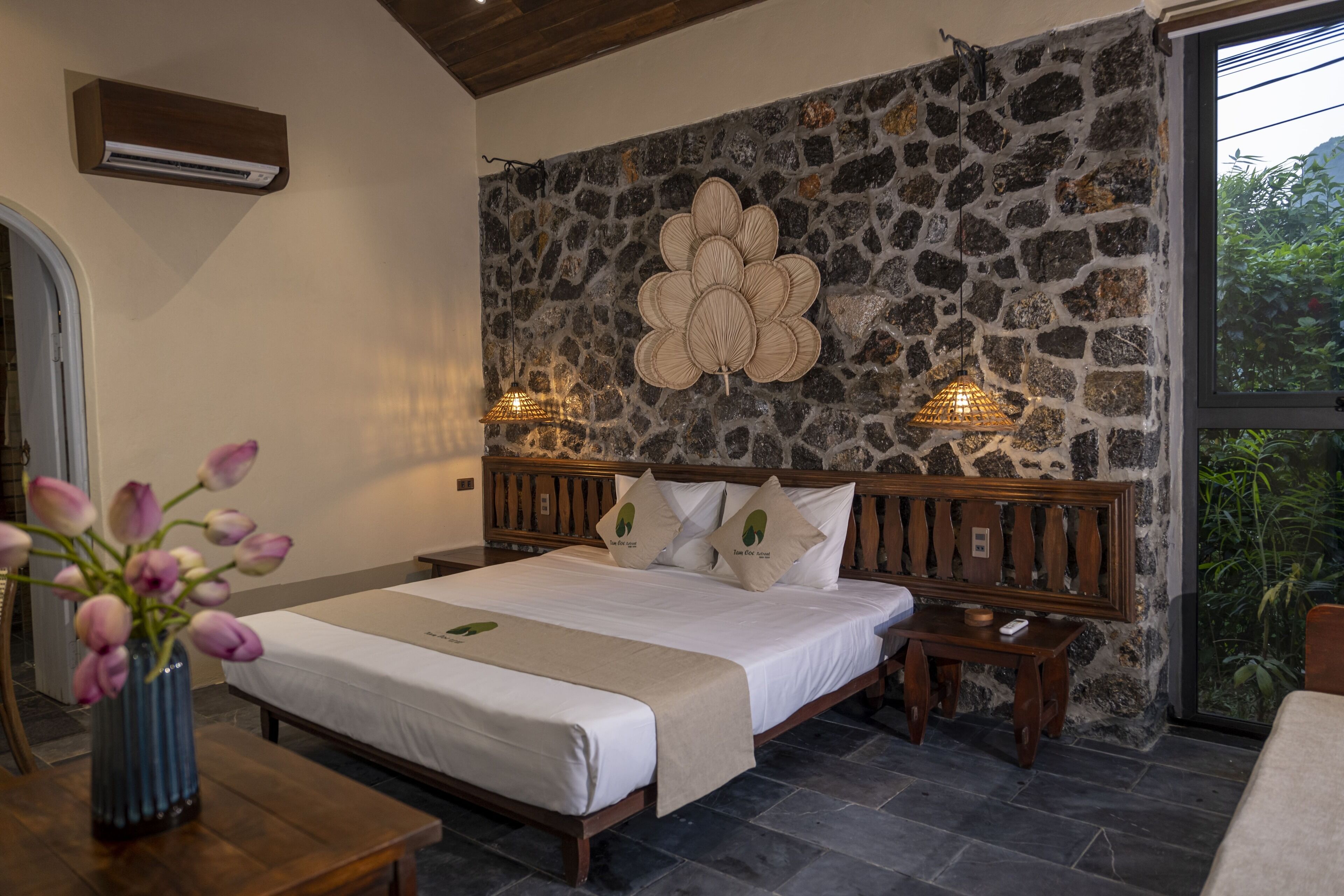 A bedroom with a stone wall, a wooden bed frame, a bedside table, and a vase of flowers.
