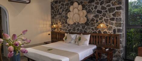 A bedroom with a stone wall, a wooden bed frame, a bedside table, and a vase of flowers.