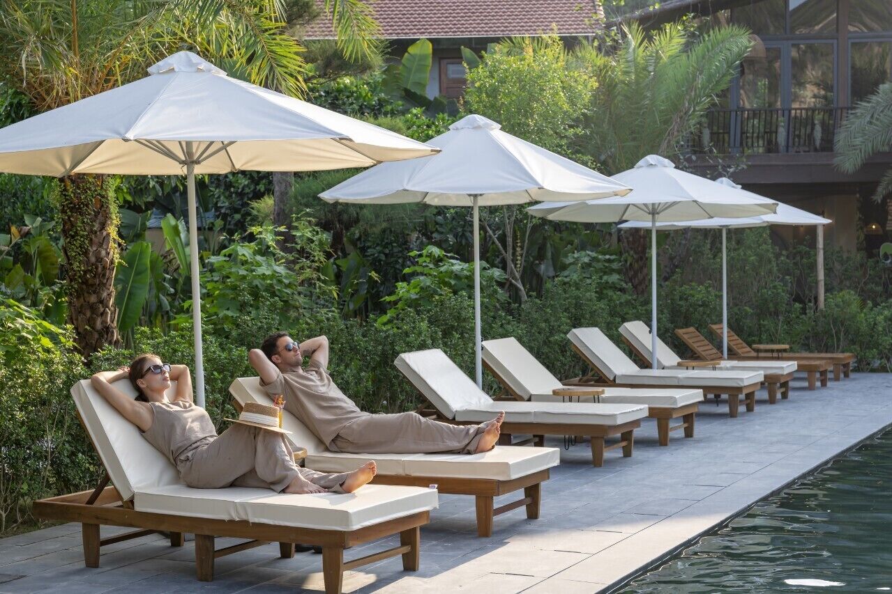 A row of sun loungers with white cushions and wooden frames, arranged along a poolside with white umbrellas providing shade.