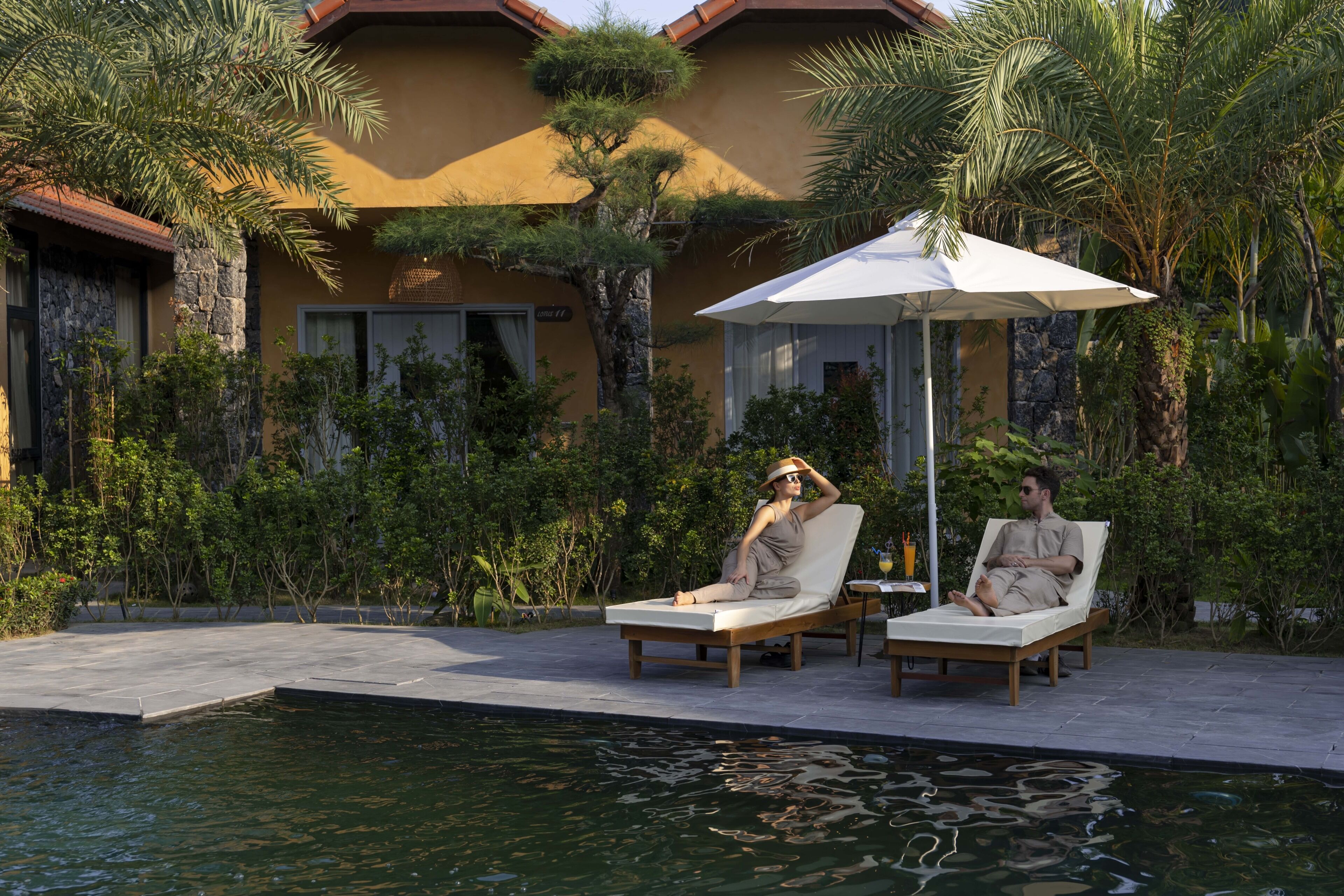 A poolside area with two lounge chairs and a large umbrella, surrounded by lush greenery and a building with a yellow facade.