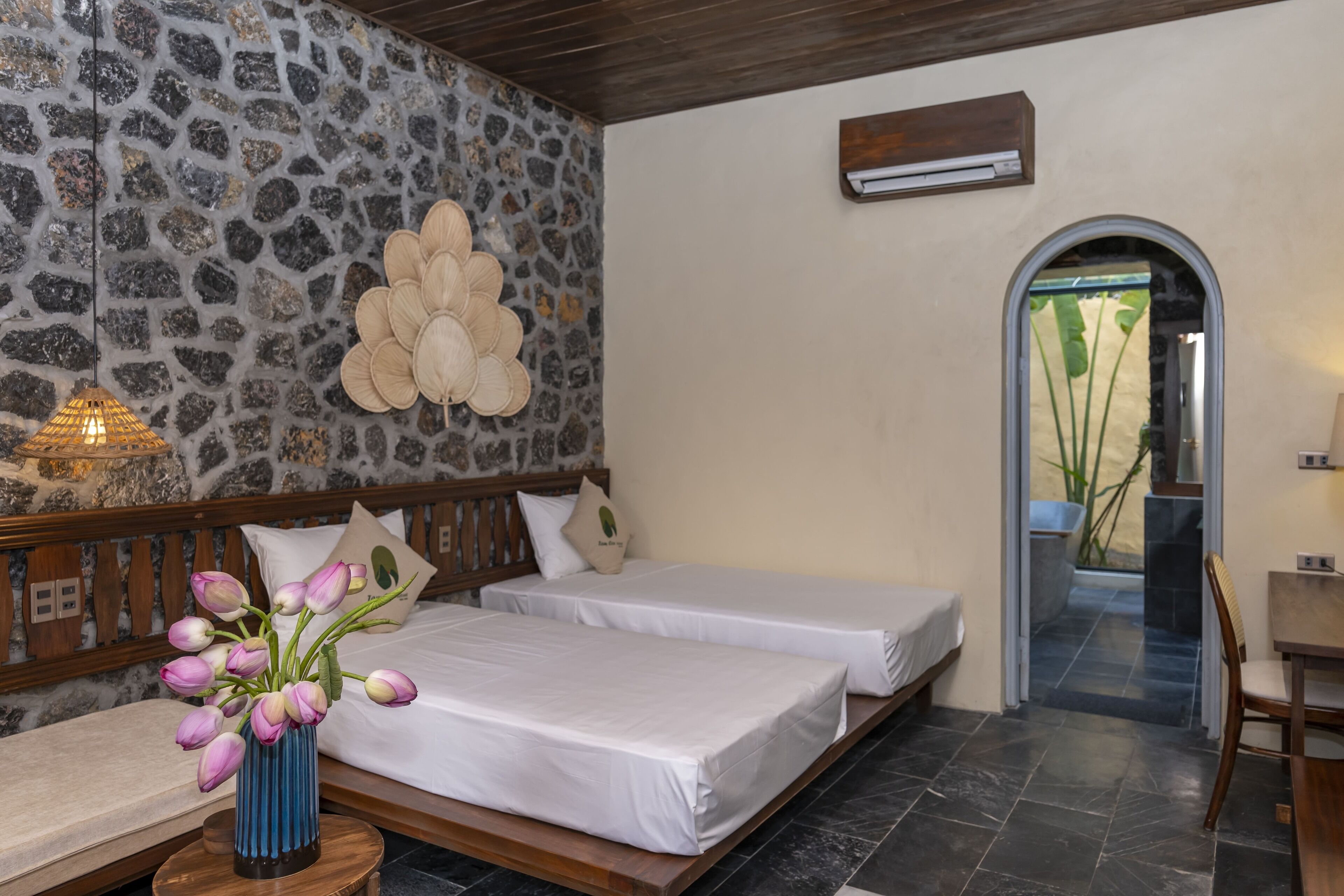 A room with two beds, a stone wall, a wooden ceiling, and a view of a garden through an arched doorway.