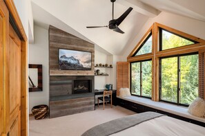Un dormitorio con una cama grande, un televisor instalado en una pared de madera, una chimenea, un escritorio con una silla y una ventana con vistas a árboles.