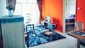 Habitació amb una paret vermella decorativa, un ventilador de sostre, zona de menjador amb taula i cadires, un televisor sobre un suport i una catifa amb disseny.