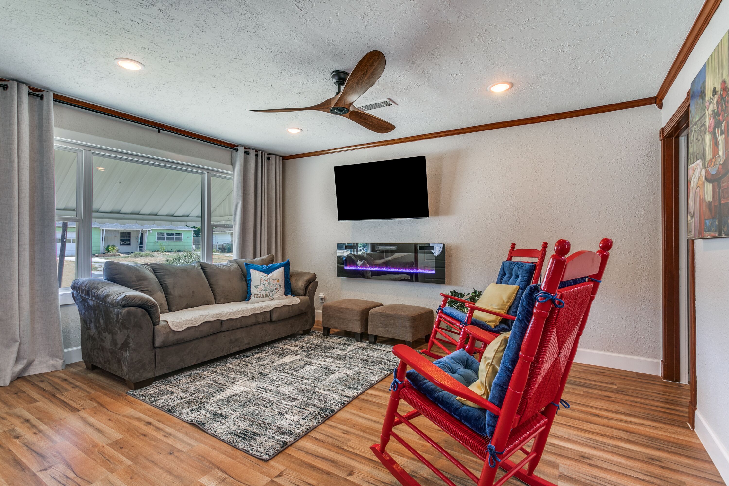 A living room with a ceiling fan, a flat-screen TV, a grey sofa, a red rocking chair, and a patterned rug.