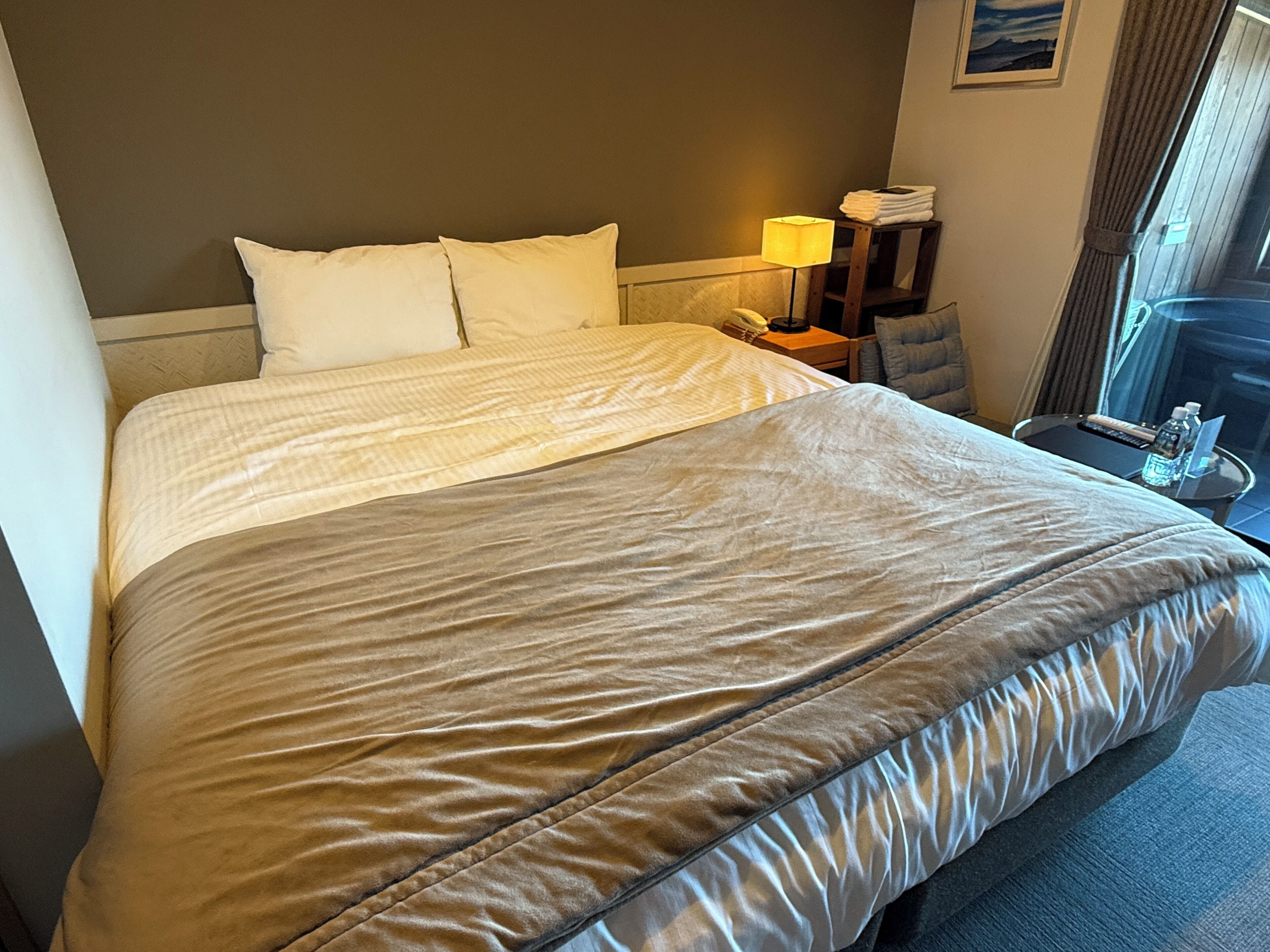 Ein Hotelzimmer mit einem großen Bett, einem Nachttisch mit Lampe, einem Sessel und einem Fenster mit Vorhängen.