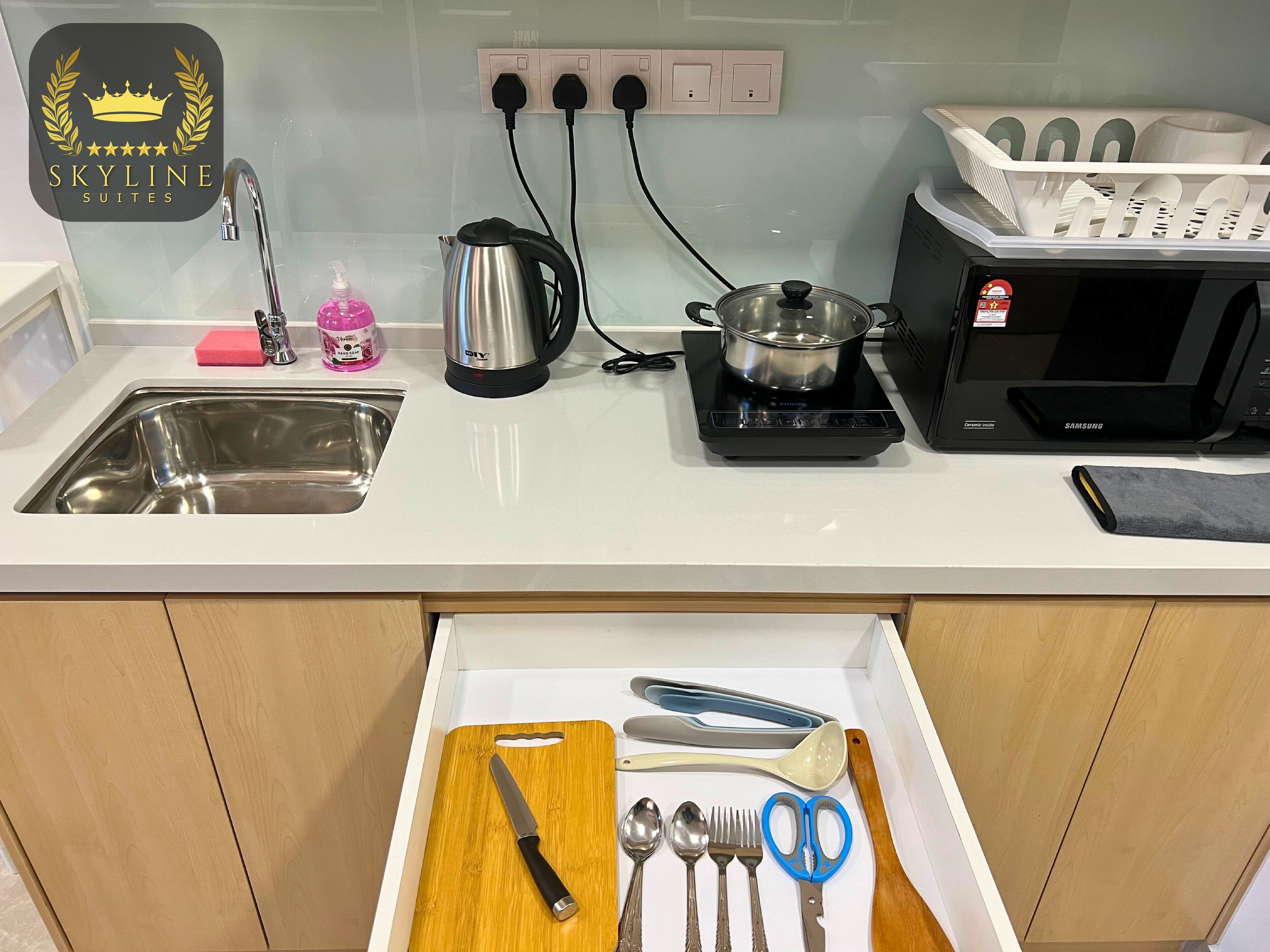 Una cocina con lavamanos, hervidor, microondas y un cajón con utensilios.