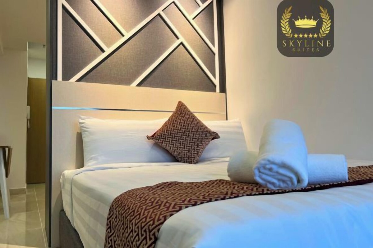 A hotel room with a neatly made bed, a patterned throw blanket, and a pillow with a geometric design.