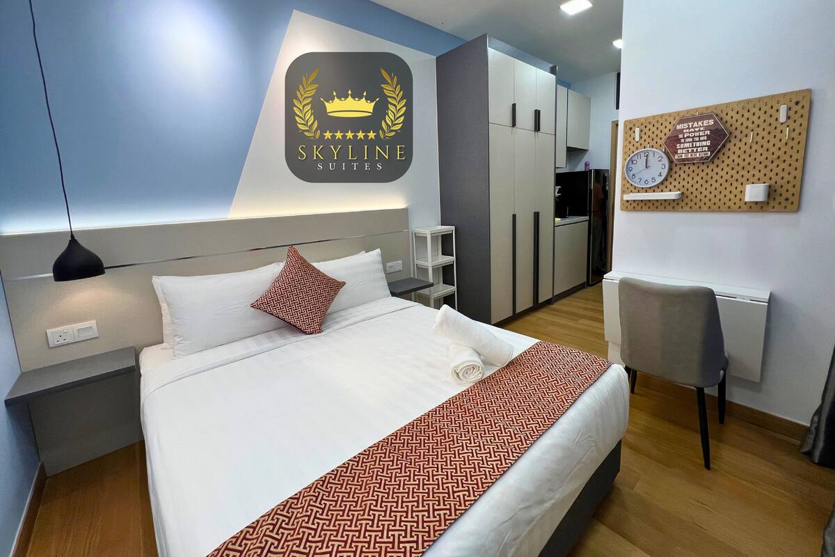 Une chambre d’hôtel équipée d’un lit, d’un bureau, d’une chaise, d’une kitchenette et d’un mur sur lequel est affiché un panneau indiquant « SKYLINE SUITES ».
