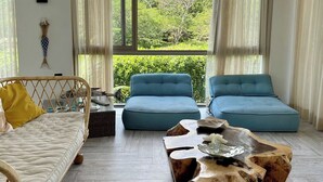 Ein geräumiges Wohnzimmer mit großem Fenster und Blick auf Grünflächen, einem blauen Sofagarnitur, einem Holztisch und einem Korbstuhl.