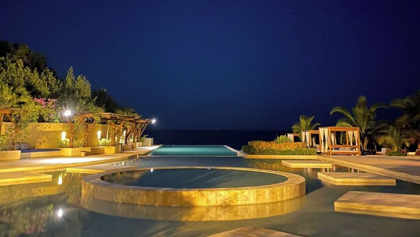 Ein Poolbereich eines Resorts bei Nacht mit mehreren Pools und Liegebereichen, umgeben von Vegetation und Blick auf den Ozean.