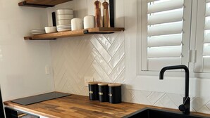 Une cuisine moderne avec des meubles blancs, un plan de travail en bois et un évier noir. Des étagères au-dessus du plan de travail servent à ranger la vaisselle et les bocaux.