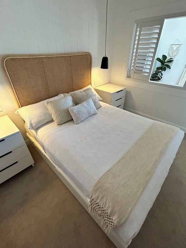 A neatly made bed with a textured white duvet and two pillows, a bedside table with a lamp, and a window with blinds and a plant. - Premium Suite Ocean Views
(Lake Cathie)