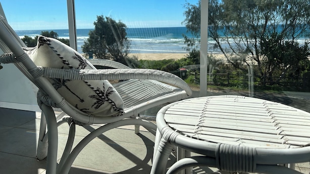 Un balcon doté de mobilier en osier, comprenant une chaise et une petite table, qui offre une vue sur une plage et l’océan.