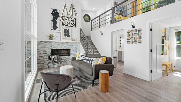 A spacious living room with a grey sofa, a black chair, a wooden stool, and a fireplace. A staircase leads to an upper level with a balcony.
