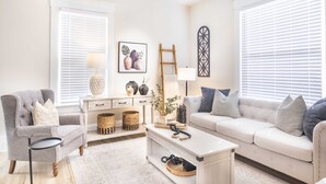 Una sala de estar con un sillón gris, un sofá blanco, una mesa de centro y una mesita auxiliar. Hay dos ventanas con persianas, un cuadro en la pared y una escalera apoyada contra la pared.