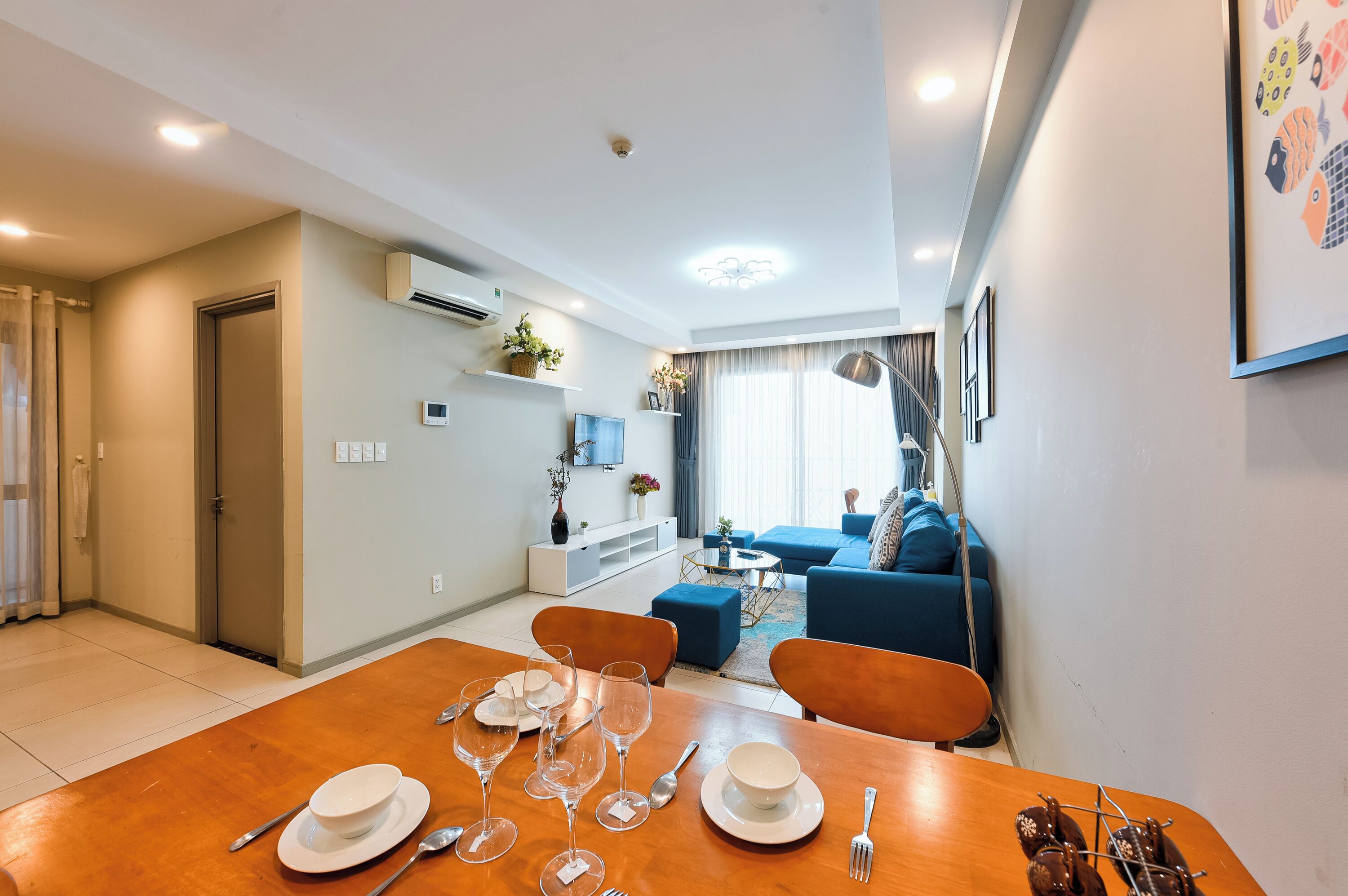 A modern living room with a blue sofa, a dining area with a wooden table set for four, and a kitchen area with a white cabinet and a hanging plant.