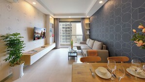 A modern living room with a sofa, a dining table set for four, a TV mounted on the wall, and a large window with curtains. - Gold View Saigon Riverside (Ho Chi Minh City)