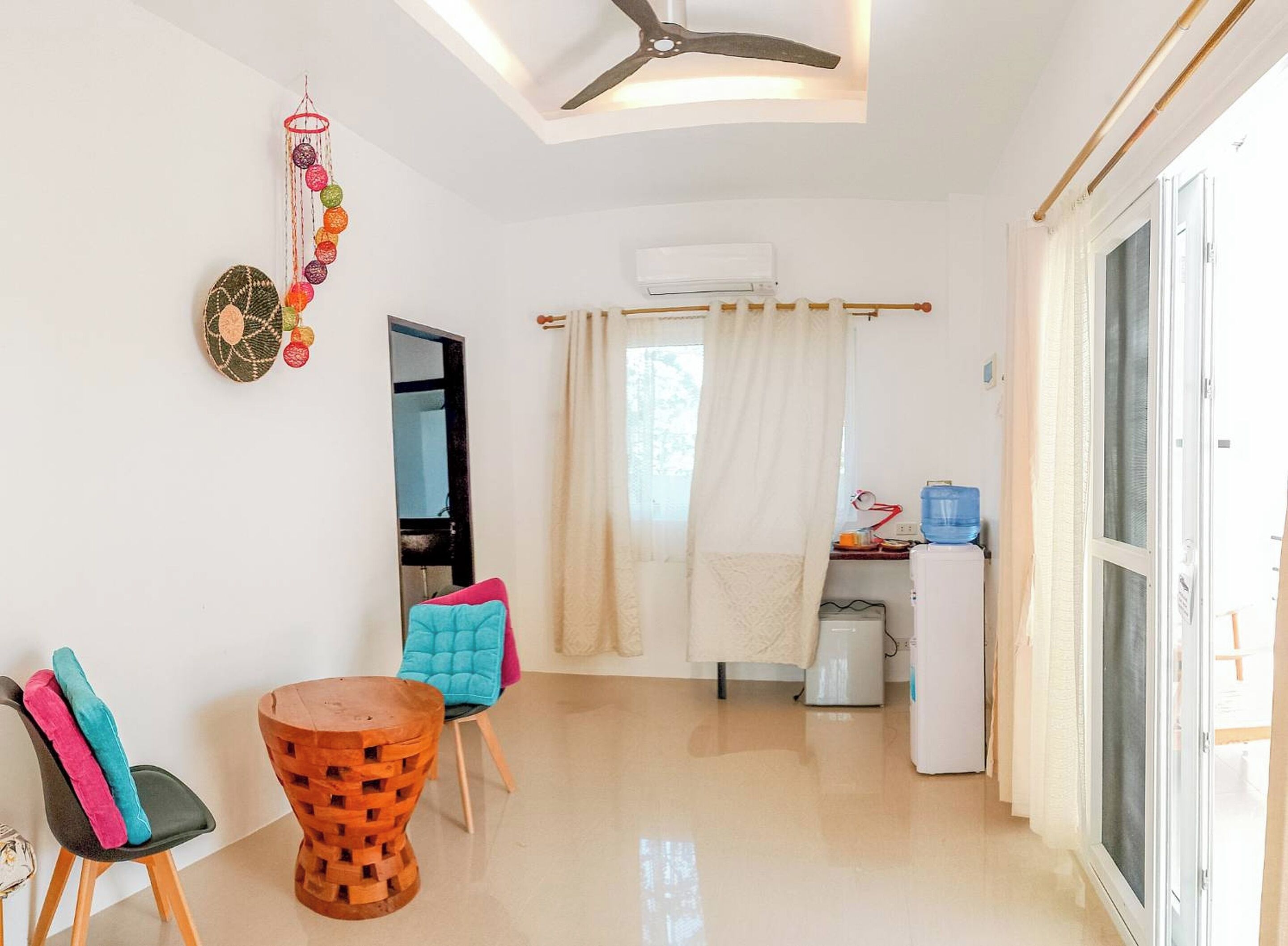 Een moderne woonkamer met een eethoek, een plafondventilator, een wandairconditioner en een keuken met een koelkast en waterdispenser.