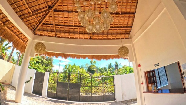 A covered outdoor area with a thatched roof, hanging lanterns, and a gate leading to a building.