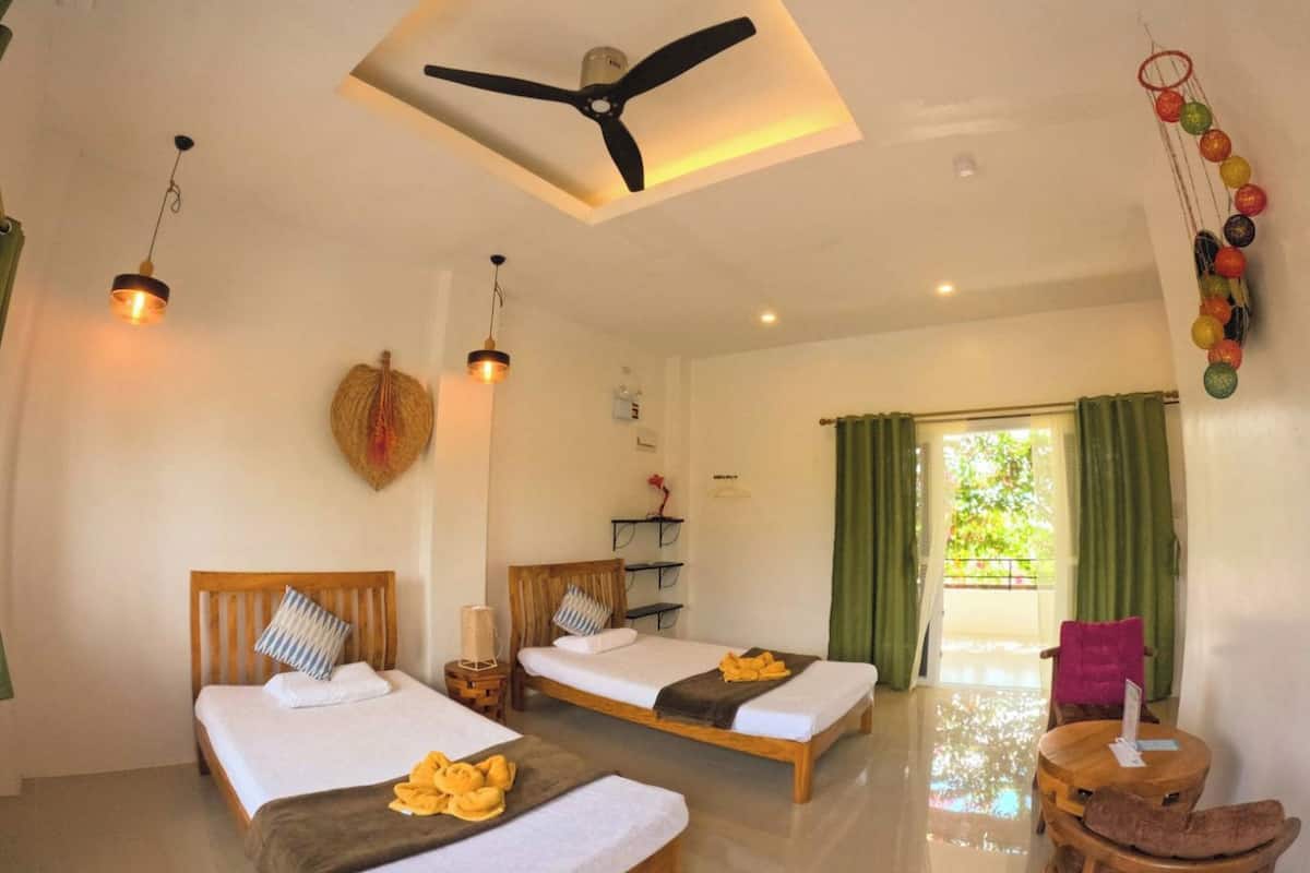 Una habitación de hotel con dos camas, un ventilador de techo, una mesita pequeña y vistas al exterior.
