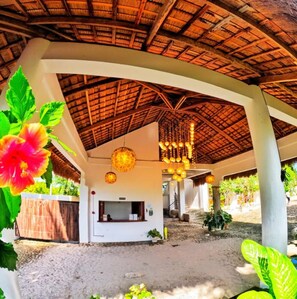 A covered outdoor area with a thatched roof, hanging light fixtures, and a white building with a window. - Divers Lodge Plumeria (Moalboal)
