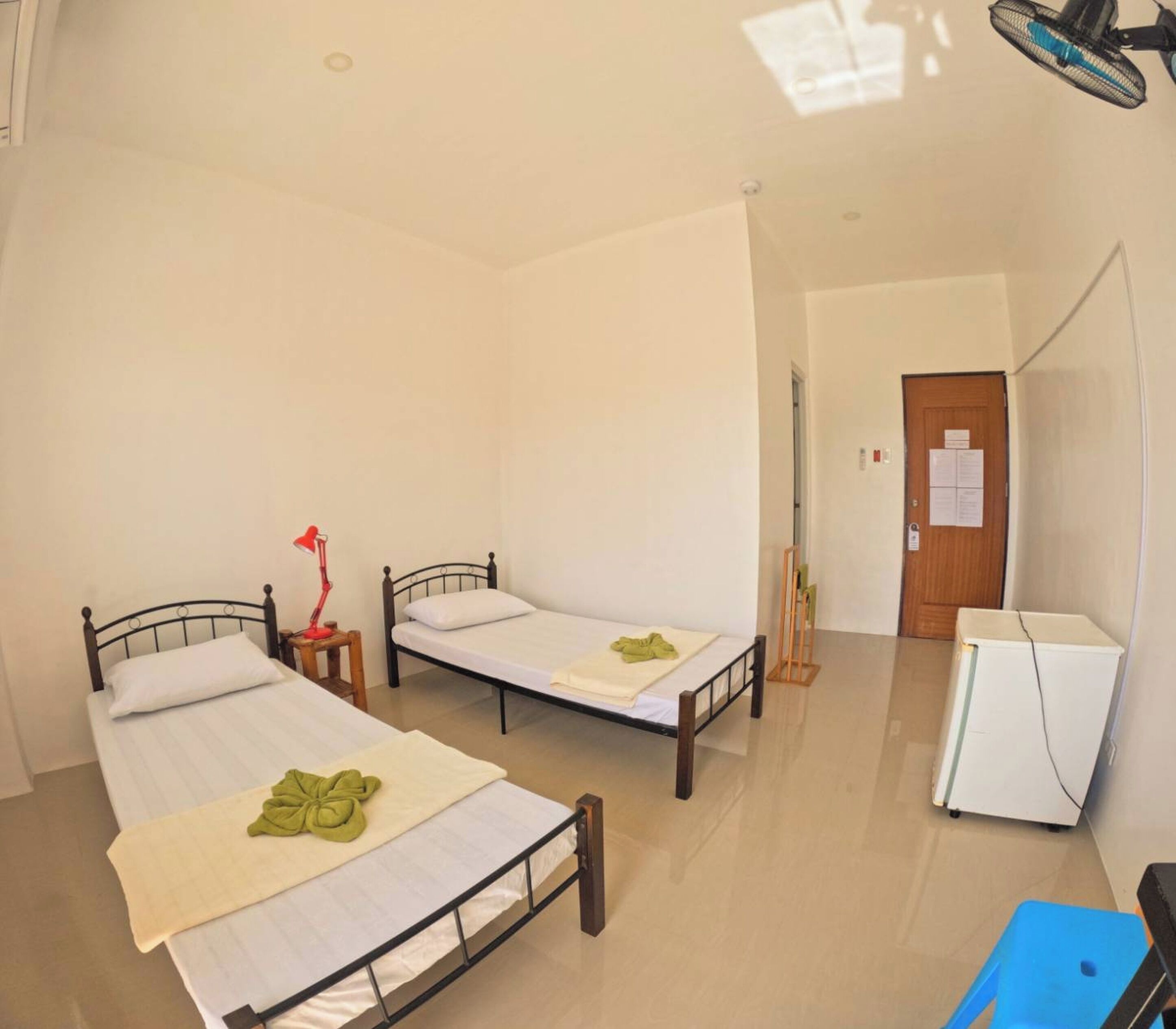 A hotel room with two single beds, a desk, a chair, a small table with a lamp, and a door with a notice.