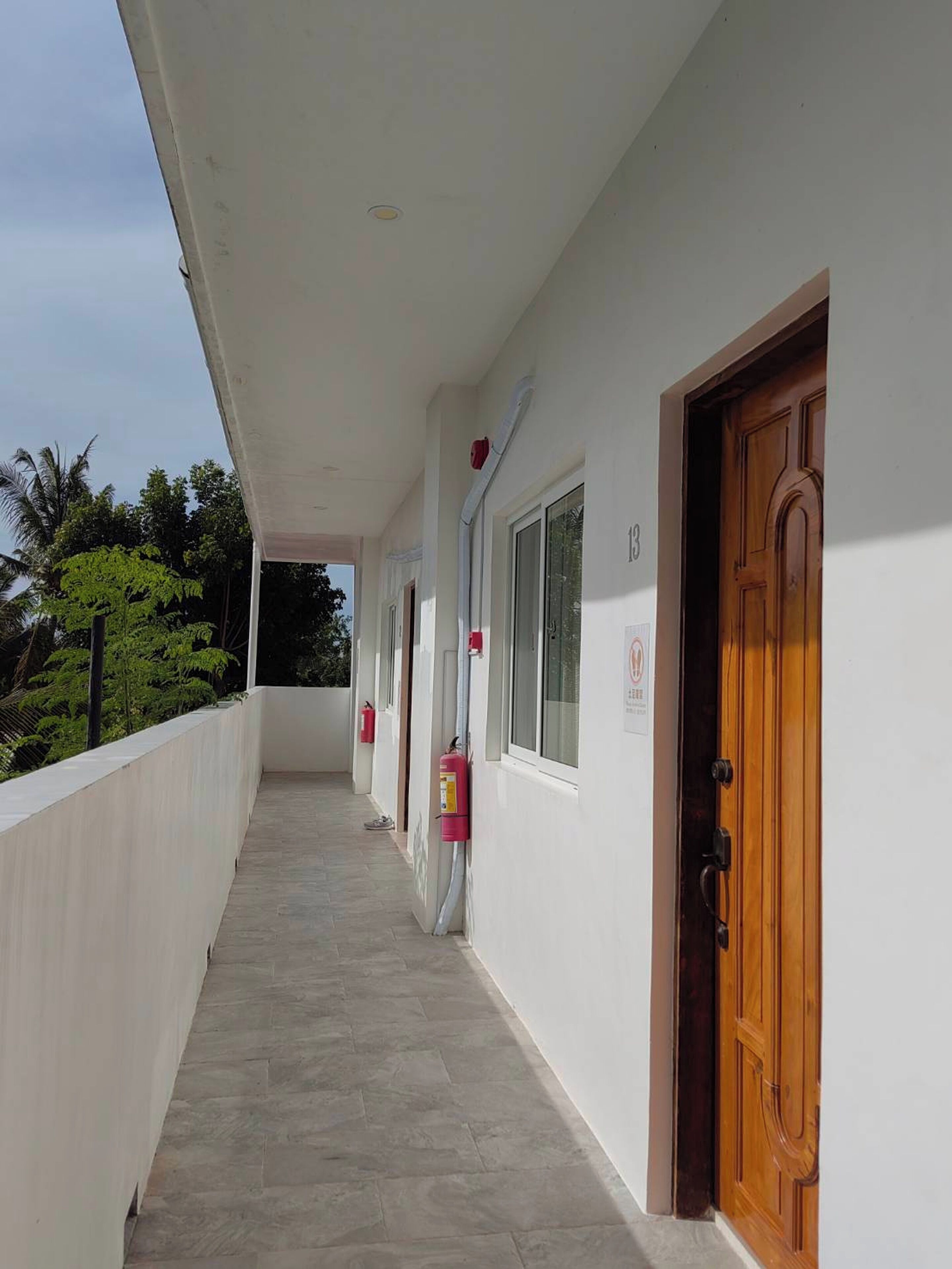 A covered corridor with a tiled floor, a wooden door on the right, and a fire extinguisher mounted on the wall.
