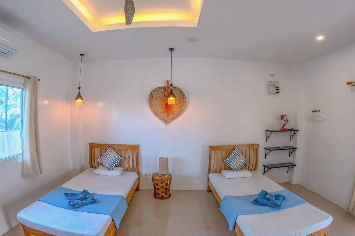 Habitación de hotel con dos camas, una lámpara de pared con forma de corazón, una mesita auxiliar y una ventana con cortinas.