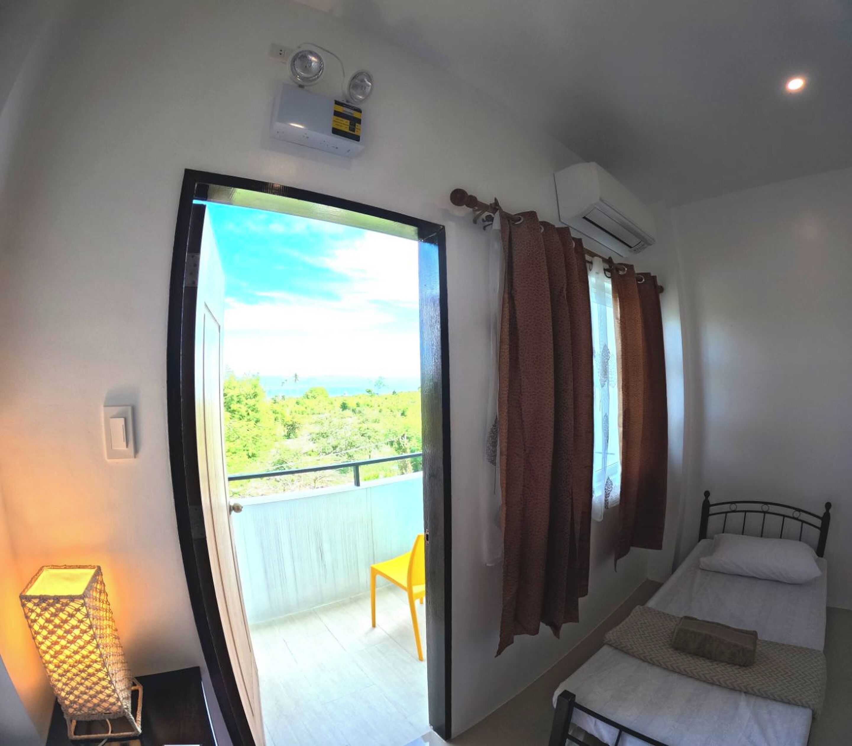 A bedroom with a bed, a chair, a balcony with a view, and an air conditioning unit.