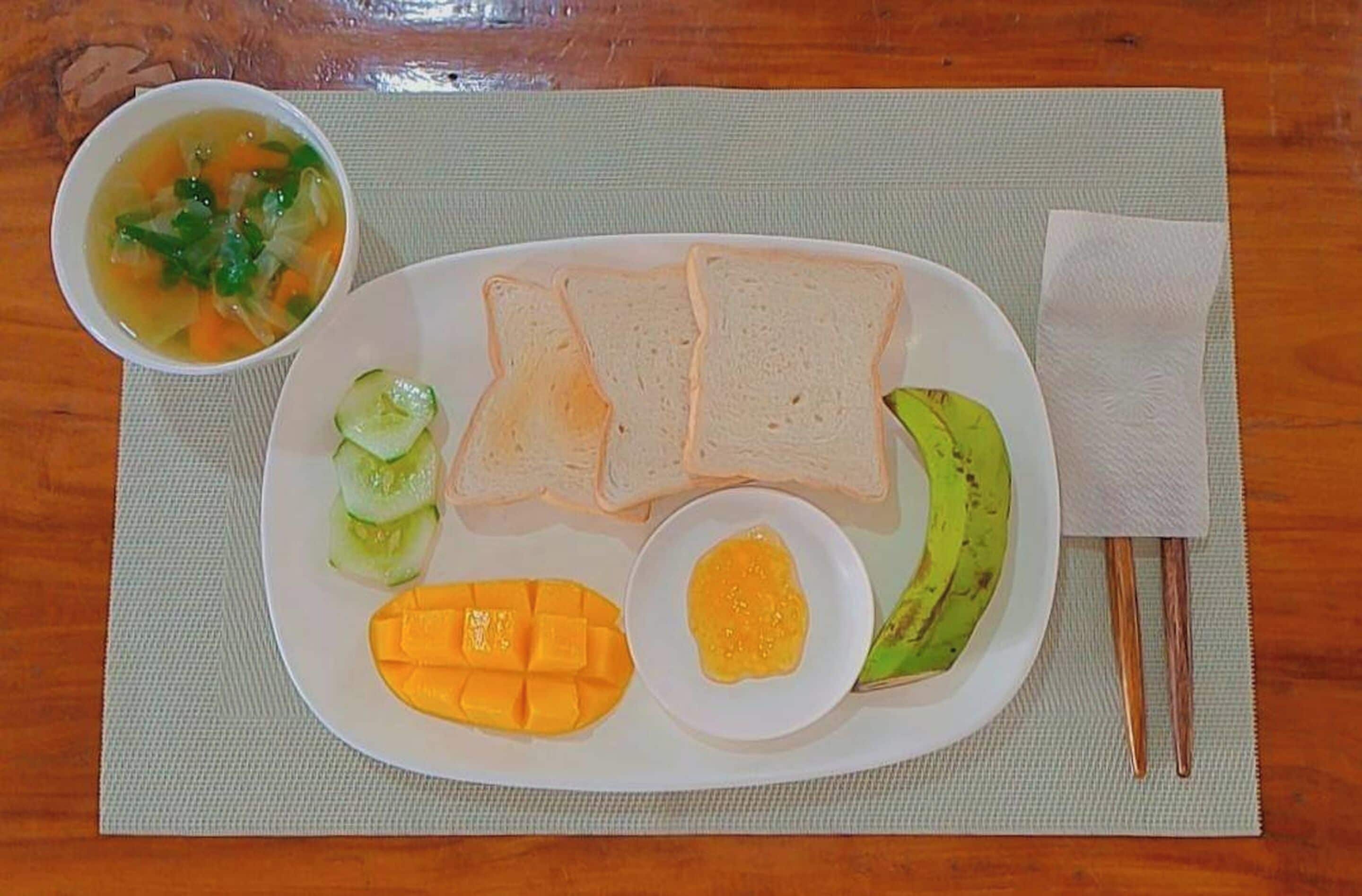 A plate with slices of bread, mango, cucumber, and a bowl of soup on a placemat.