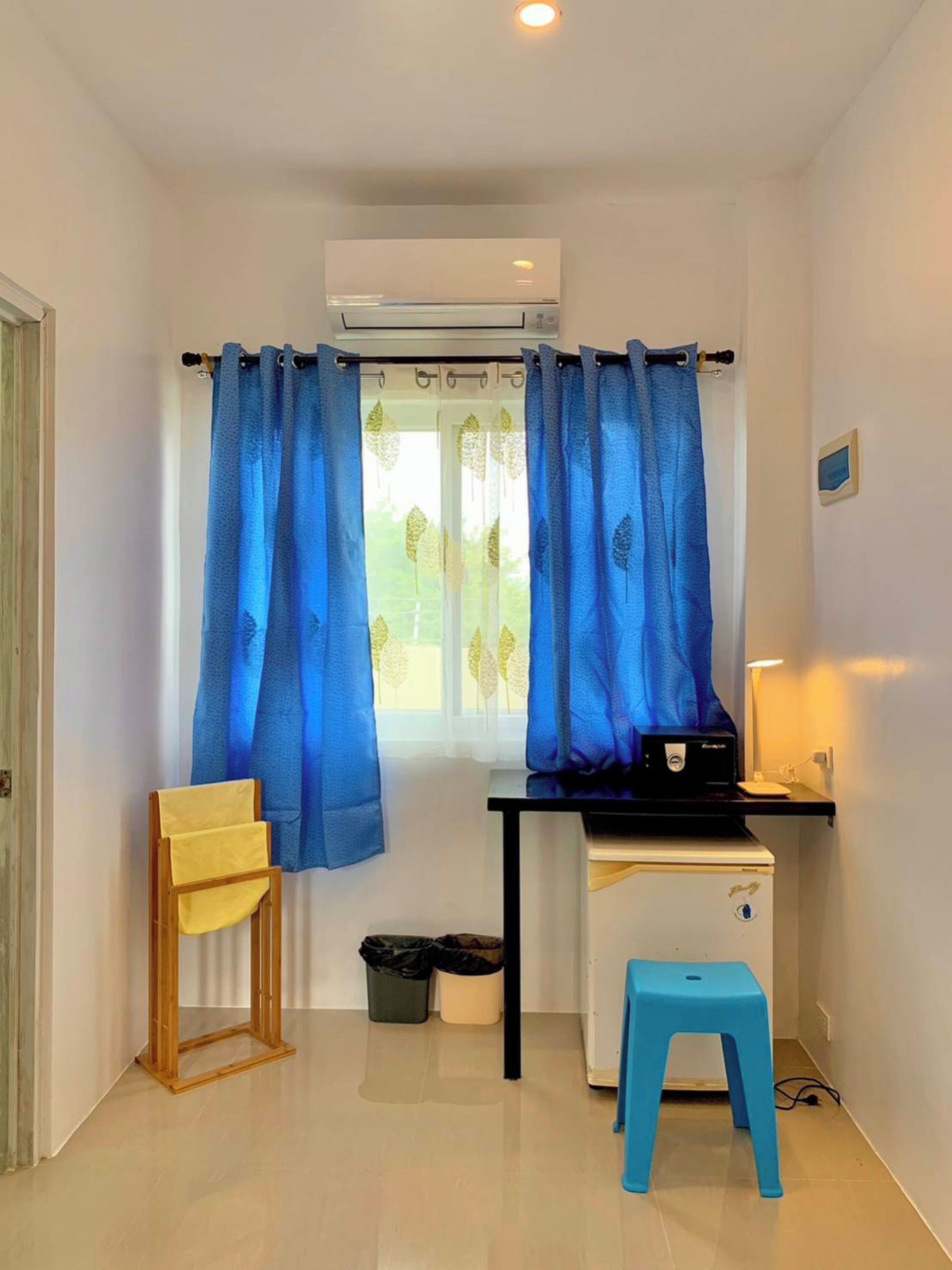A small room with a desk, a chair, a stool, and a window with blue curtains.