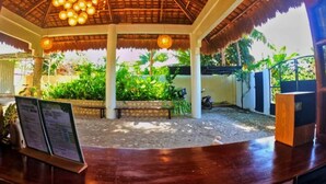 A covered outdoor seating area with a wooden bench and a thatched roof, surrounded by greenery.