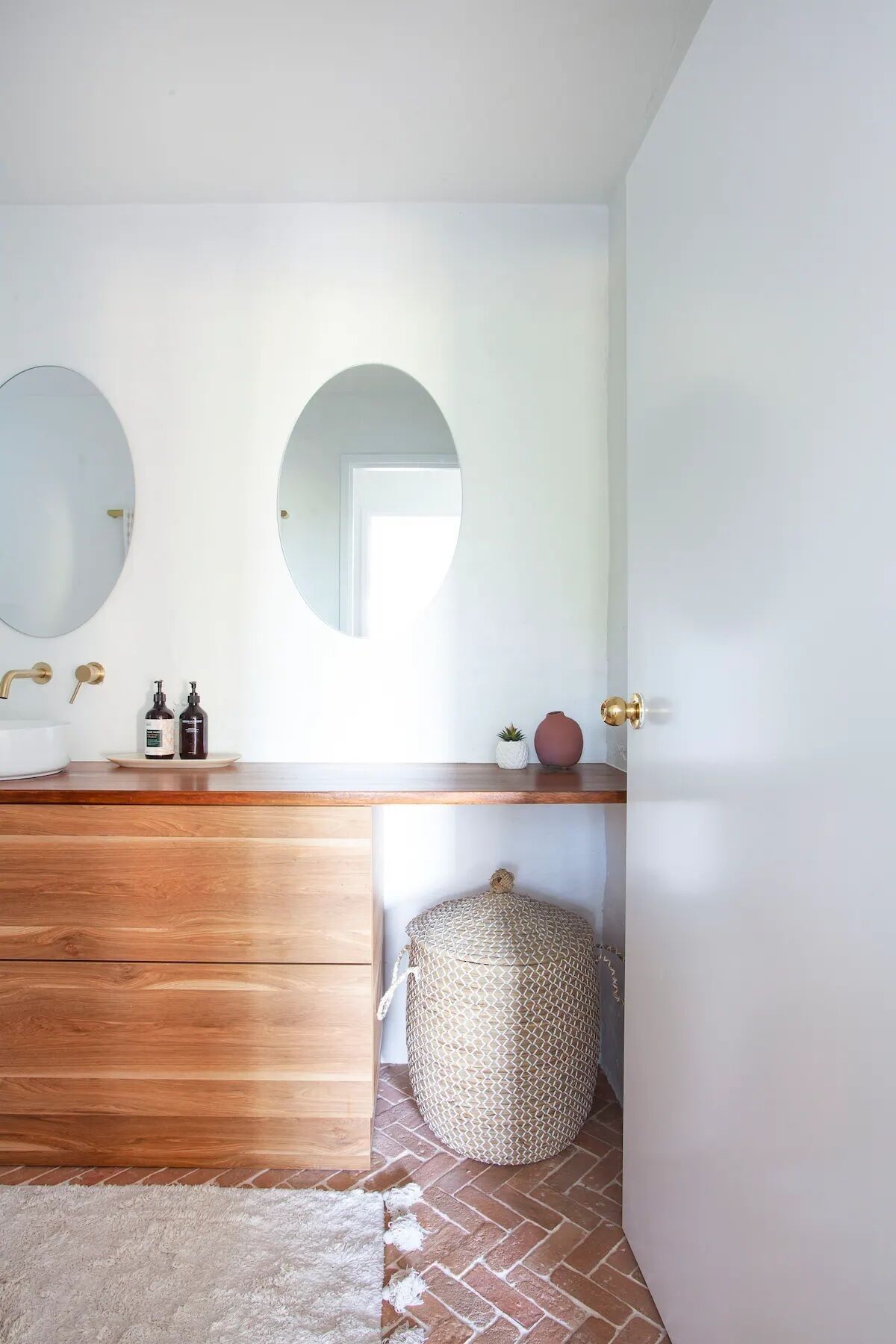 Un bagno con un mobile vanitario in legno, due specchi rotondi, un lavandino bianco e uno sgabello rotondo in fibra naturale.