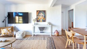 Ein modernes Wohnzimmer mit Fernseher, einem Gemälde, einem Konsolentisch, einem Essbereich mit Holztisch und Stühlen sowie einem gemusterten Teppich.