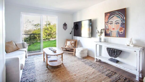 Ein modernes Wohnzimmer mit einer Couch, einem runden Tisch, einer weißen Kommode mit Dekorationen, einem großen Gemälde und einem an der Wand befestigten Fernseher.