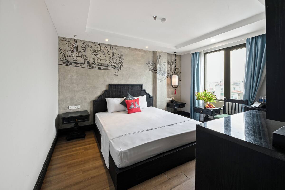 A modern hotel room with a large bed, a small desk, a nightstand, a window with curtains, and a city view.