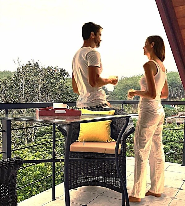 Un balcón con una silla de mimbre, una mesa con una bandeja roja y vistas al verdor.