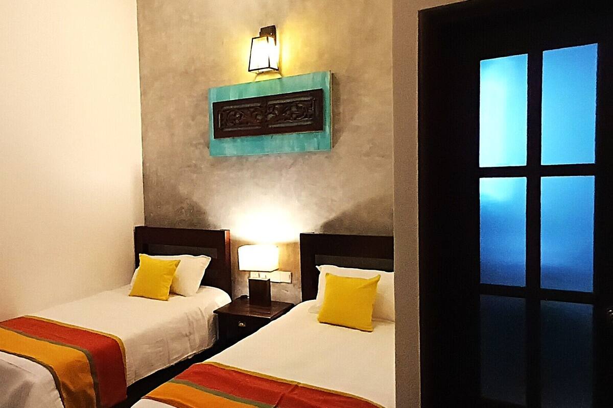A hotel room with two single beds, each with a white bedspread and a colorful striped blanket. There is a nightstand with a lamp between the beds, and a wall-mounted light fixture above the bed.