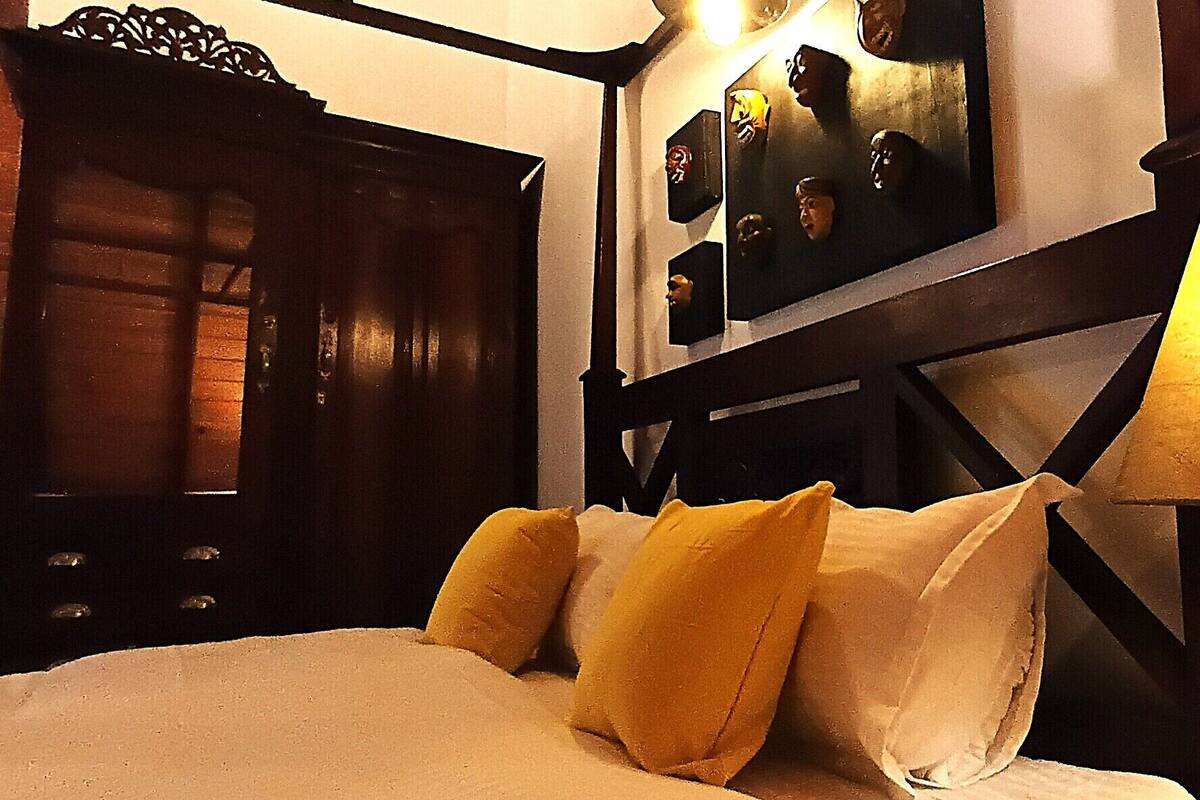 A bed with white and yellow pillows, a wooden headboard, a wall-mounted lamp, and a mirror with framed pictures.