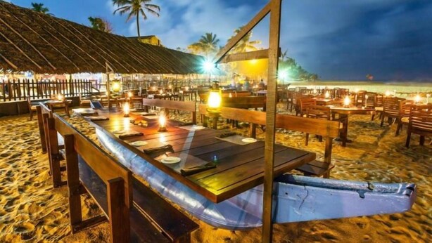 Ein Restaurant direkt am Strand mit Terrassenplätzen, Holztischen und einem Restaurantgebäude mit Reetdach.
