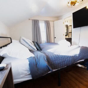 Una cama bien hecha con edredón azul y blanco, una mesita de noche de madera y un televisor de pantalla plana montado en la pared.