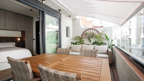 A modern room with a wooden dining table, wicker chairs, a sofa with cushions, and a large window offering a view of the city.