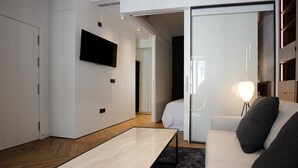 A modern hotel room with a flat-screen TV mounted on the wall, a sofa with cushions, a small table, and a bedside lamp.