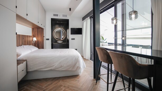 A modern bedroom with a large bed, a wooden headboard, a circular mirror, a TV, and a view of the outdoors through large windows.