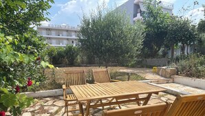 Een terras buiten met een houten tafel en stoelen, omgeven door groen en een stenen pad.