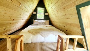 A cozy attic bedroom with wooden walls, a bed with a fluffy blanket, and a window with a view of greenery.